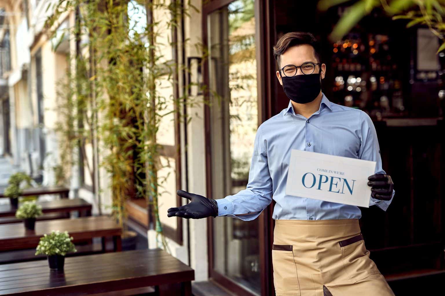 Plan for Jobs - A waiter stands in front of a restaurant with a sign that reads 'Come in we're Open'. The waiter is wearing a mask and gloves. The Eat out to Help Out Scheme encourages people to return to hospitality establishments