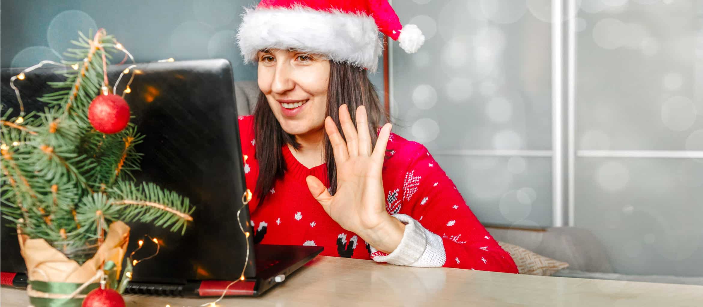 Virtual Christmas Party - employee sits in front of a laptop waving, wearing a Christmas Jumper and a Santa Hat