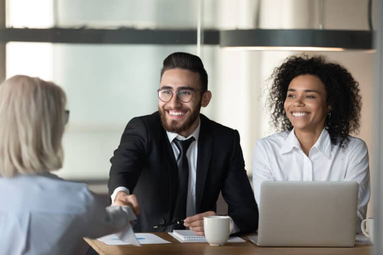 How to choose an HR outsourcing company - three people sit at a table for a meeting about outsourcing HR. On the table is a laptop, stationary and coffee mug. Two of the individuals are shaking hands