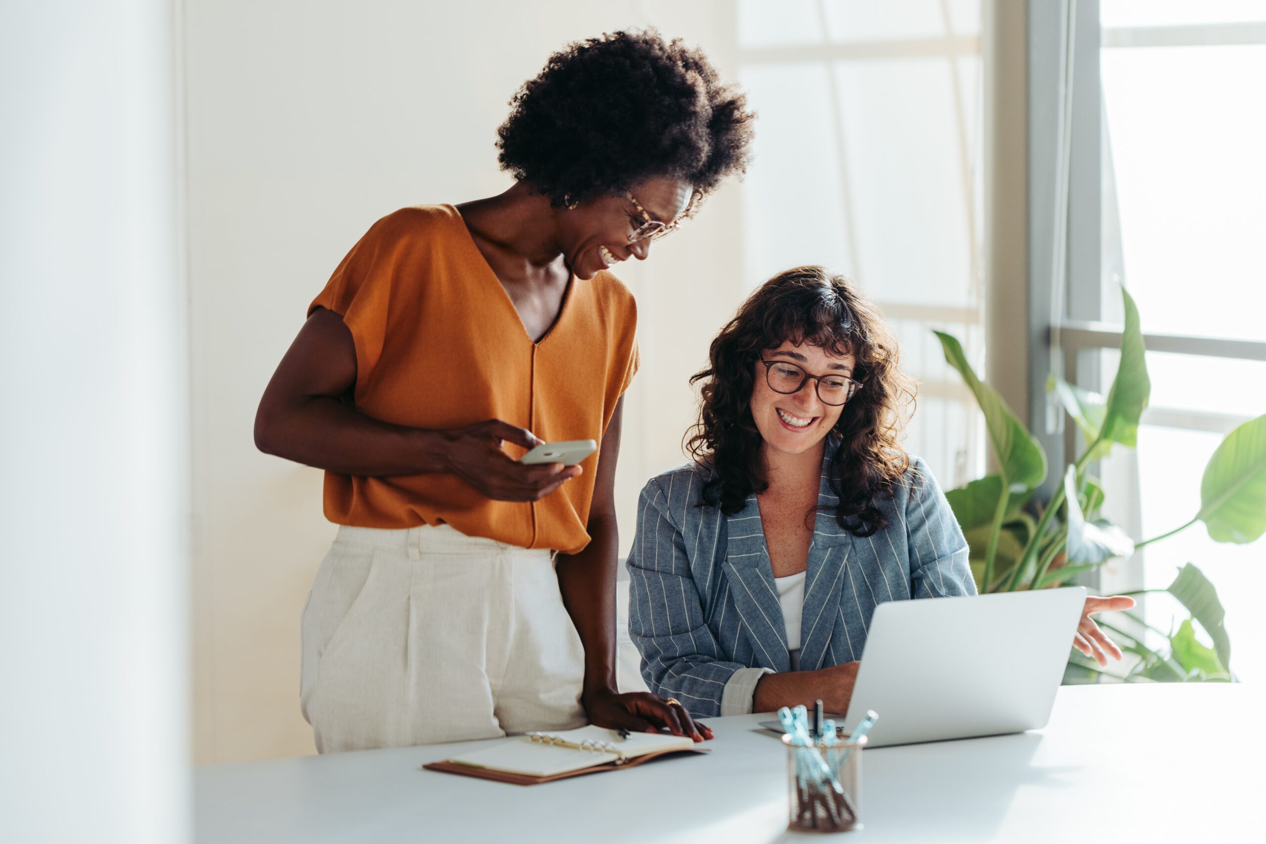 Happy,Professional,Women,Collaborating,With,Enthusiasm,In,A,Modern,Office Two women talking in front of a laptop. Ways to celebrate international women's day in your organisation.